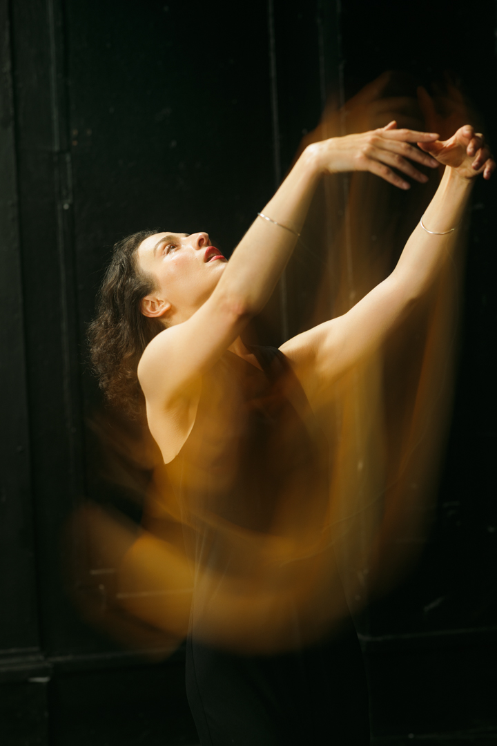 A dancer arched backwards, arms reaching upward, lit warm against a black room.